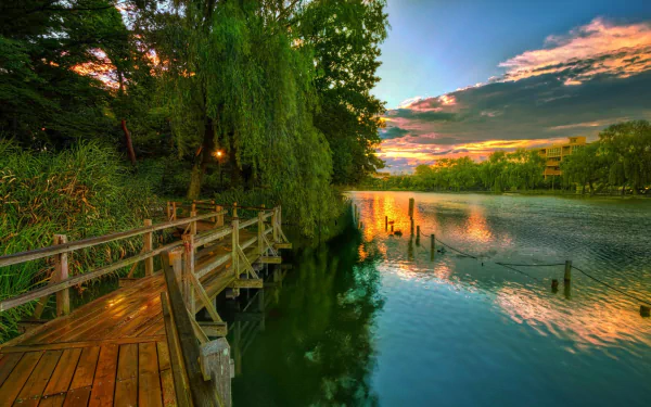 A serene boardwalk surrounded by lush green trees stretches along a calm lake under a colorful sunset sky, captured in vibrant HD photography.