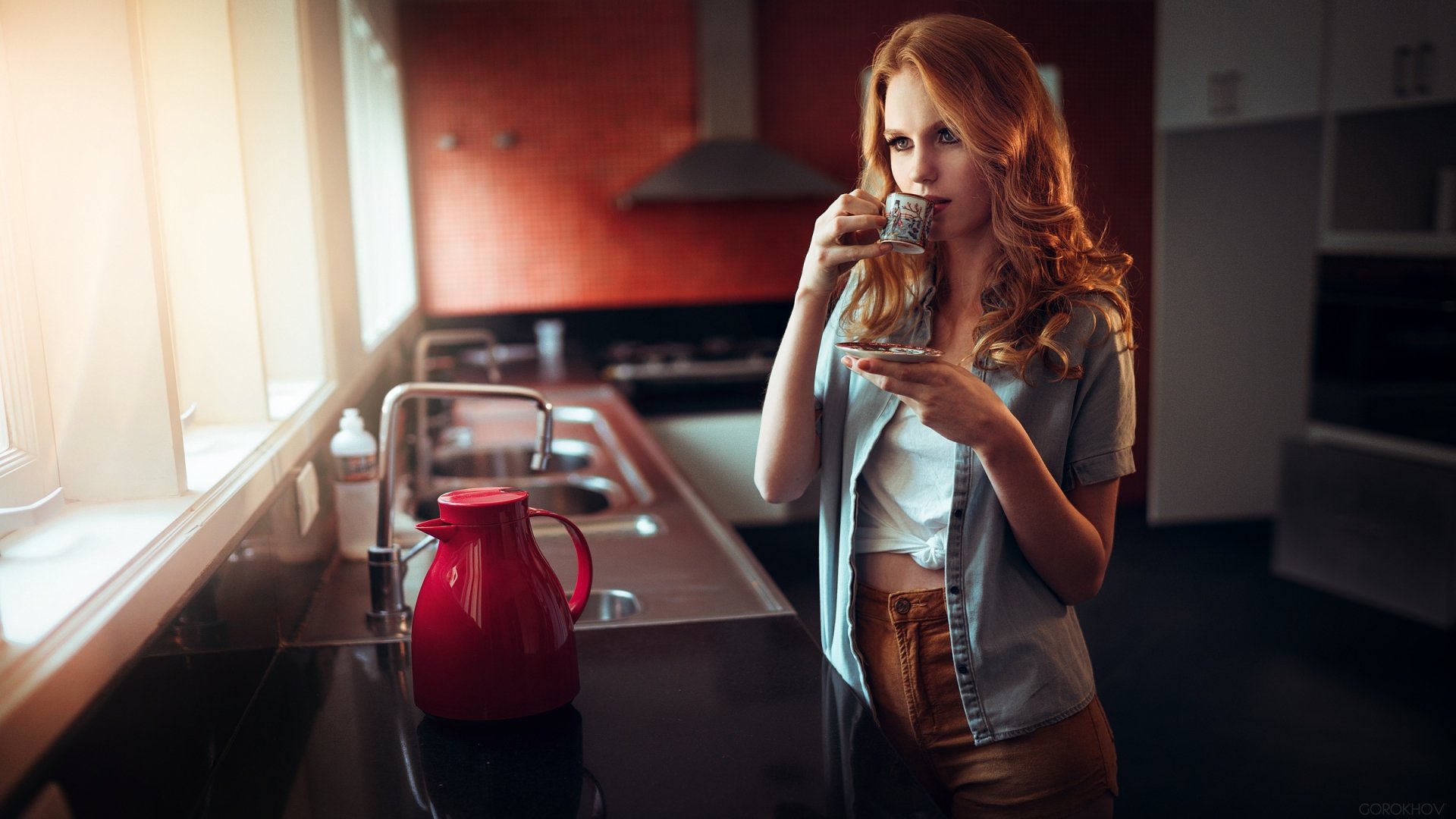 A red-headed woman stands in a modern kitchen, sipping from a small cup, with a red kettle on the counter. This HD desktop wallpaper and background highlights the kitchen's sleek design and warm lighting.