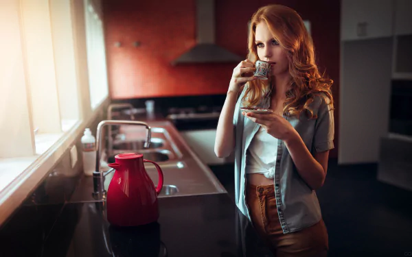 A red-headed woman stands in a modern kitchen, sipping from a small cup, with a red kettle on the counter. This HD desktop wallpaper and background highlights the kitchen's sleek design and warm lighting.