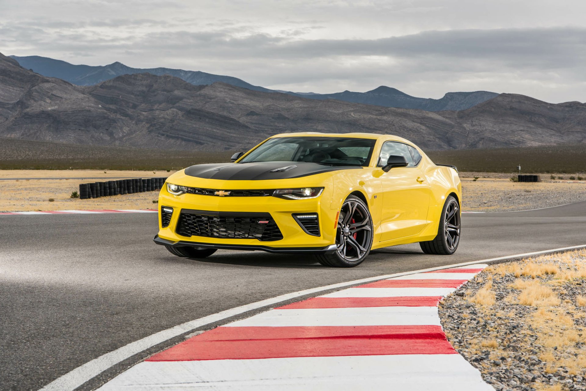 A bright yellow Chevrolet Camaro 1LE muscle car speeding on a racetrack set against a mountainous backdrop, captured in a high-definition desktop wallpaper.