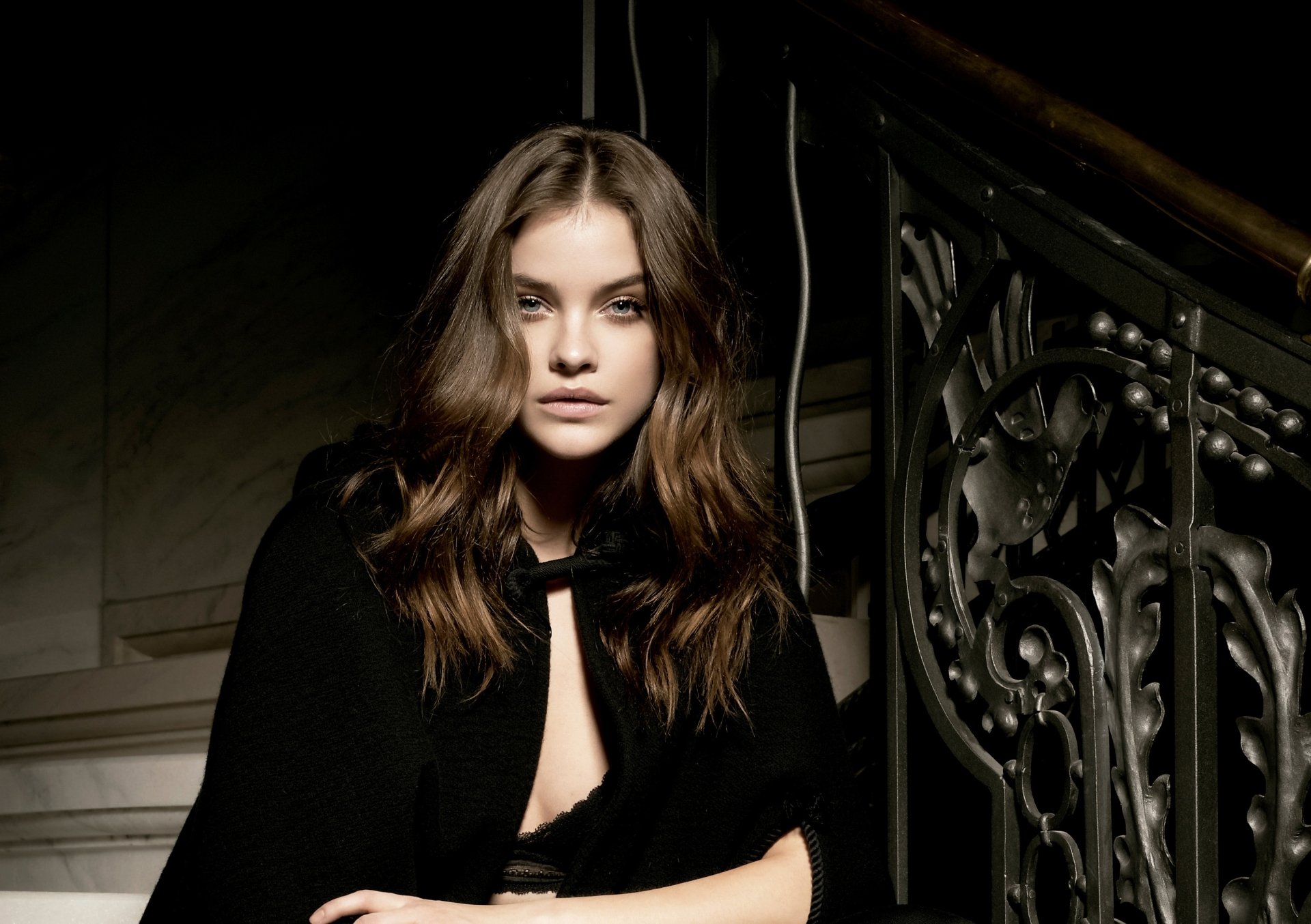HD wallpaper featuring Barbara Palvin, Hungarian brunette model with blue eyes, posing beside an ornate wrought-iron staircase in a dimly lit setting.