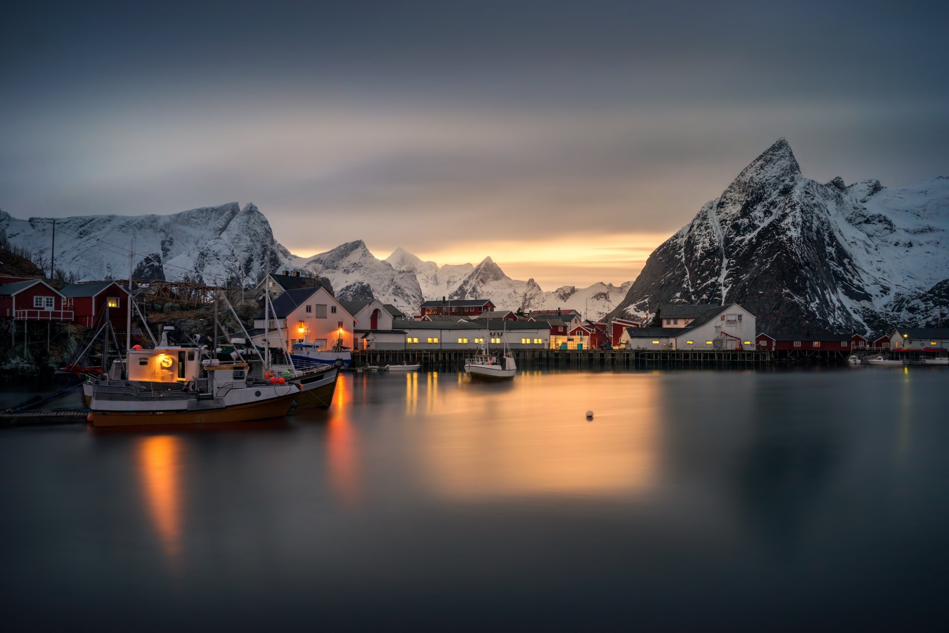 HD desktop wallpaper showcasing a serene Lofoten village with boats docked on calm water, surrounded by snowy mountains under a twilight sky. Photography capturing stunning natural beauty and peaceful evening ambiance.