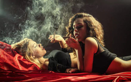 Two women, one brunette and one blonde, are smoking cigars on a red satin sheet, creating a moody, intimate atmosphere. This HD desktop wallpaper and background highlights their relaxed demeanor.