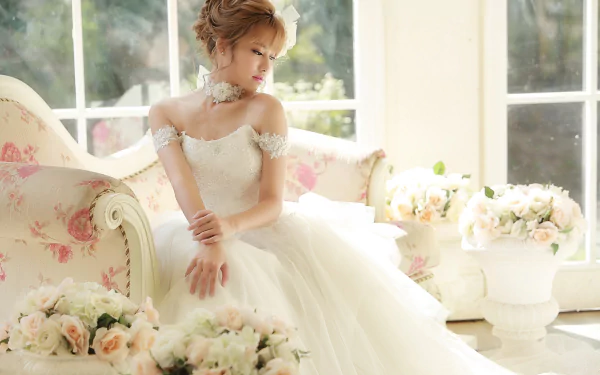 A bride with brunette hair in a white wedding dress sits on a floral couch, surrounded by bouquets of flowers, in a bright room with large windows.