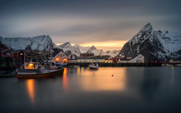 HD desktop wallpaper showcasing a serene Lofoten village with boats docked on calm water, surrounded by snowy mountains under a twilight sky. Photography capturing stunning natural beauty and peaceful evening ambiance.