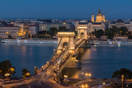 HD desktop wallpaper: nighttime view of Budapest's illuminated man-made Chain Bridge over the Danube in Hungary, with city skyline and St. Stephen's Basilica reflected on the river.