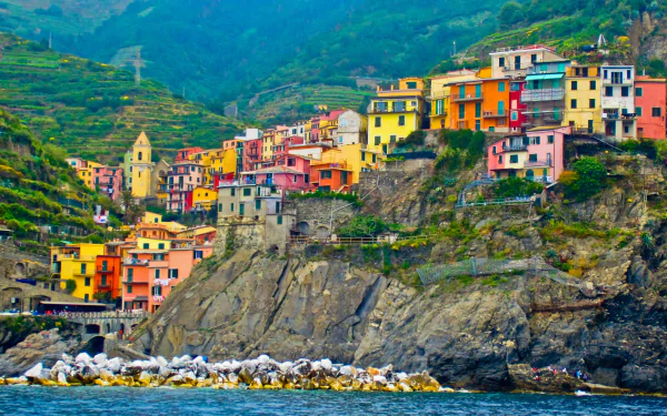 Vibrant village of Manarola in Cinque Terre, Italy, showcasing colorful houses perched on a rocky cliff with a mountain backdrop. HD desktop wallpaper.