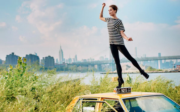 Alicia Vikander, Swedish brunette actress, poses stylishly atop a taxi with a cityscape and river in the background in this HD desktop wallpaper.