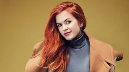 Close-up portrait of Australian actress Isla Fisher with red hair and brown eyes, wearing a camel coat and gray turtleneck against a muted yellow background.