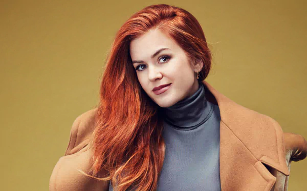 Close-up portrait of Australian actress Isla Fisher with red hair and brown eyes, wearing a camel coat and gray turtleneck against a muted yellow background.