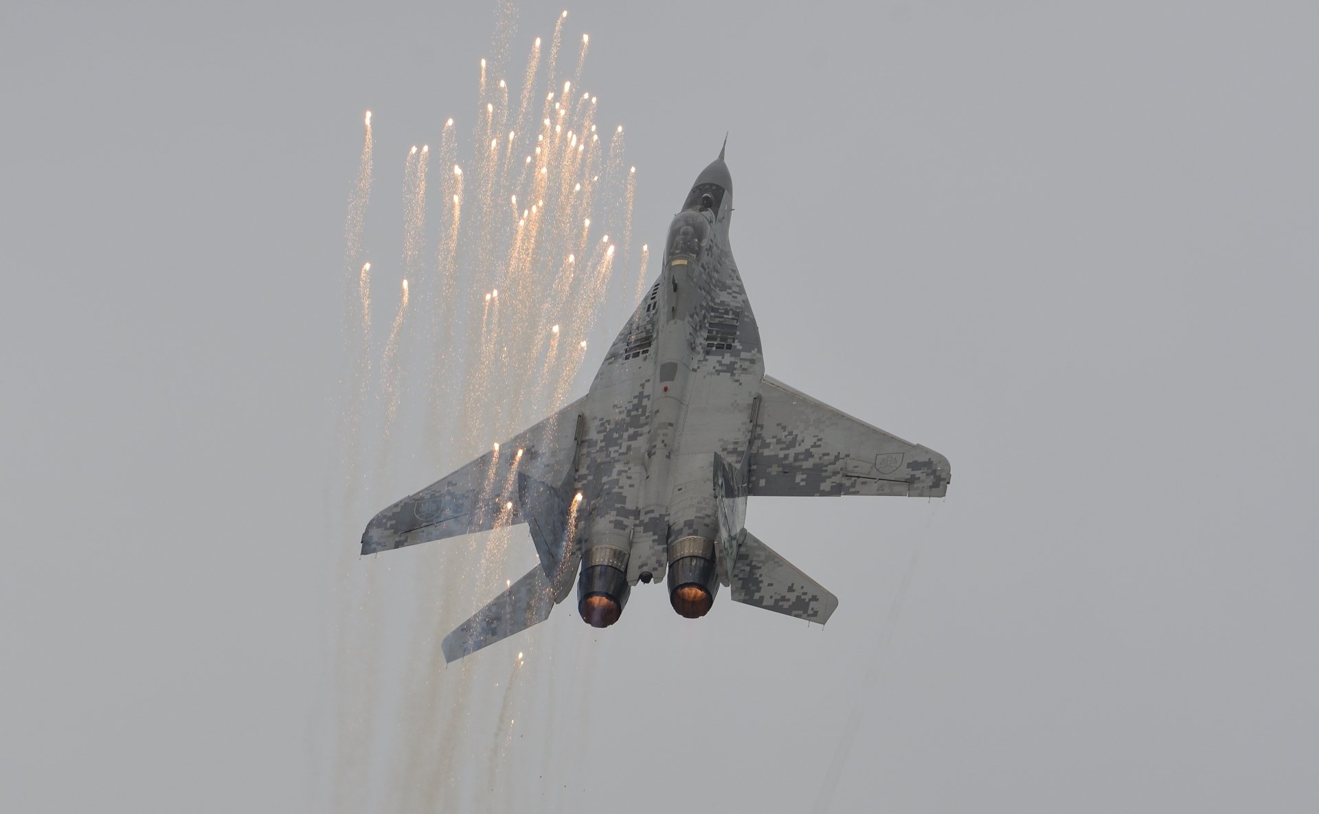 HD desktop wallpaper featuring a Mikoyan MiG-29 military jet fighter aircraft releasing flares against a gray sky.