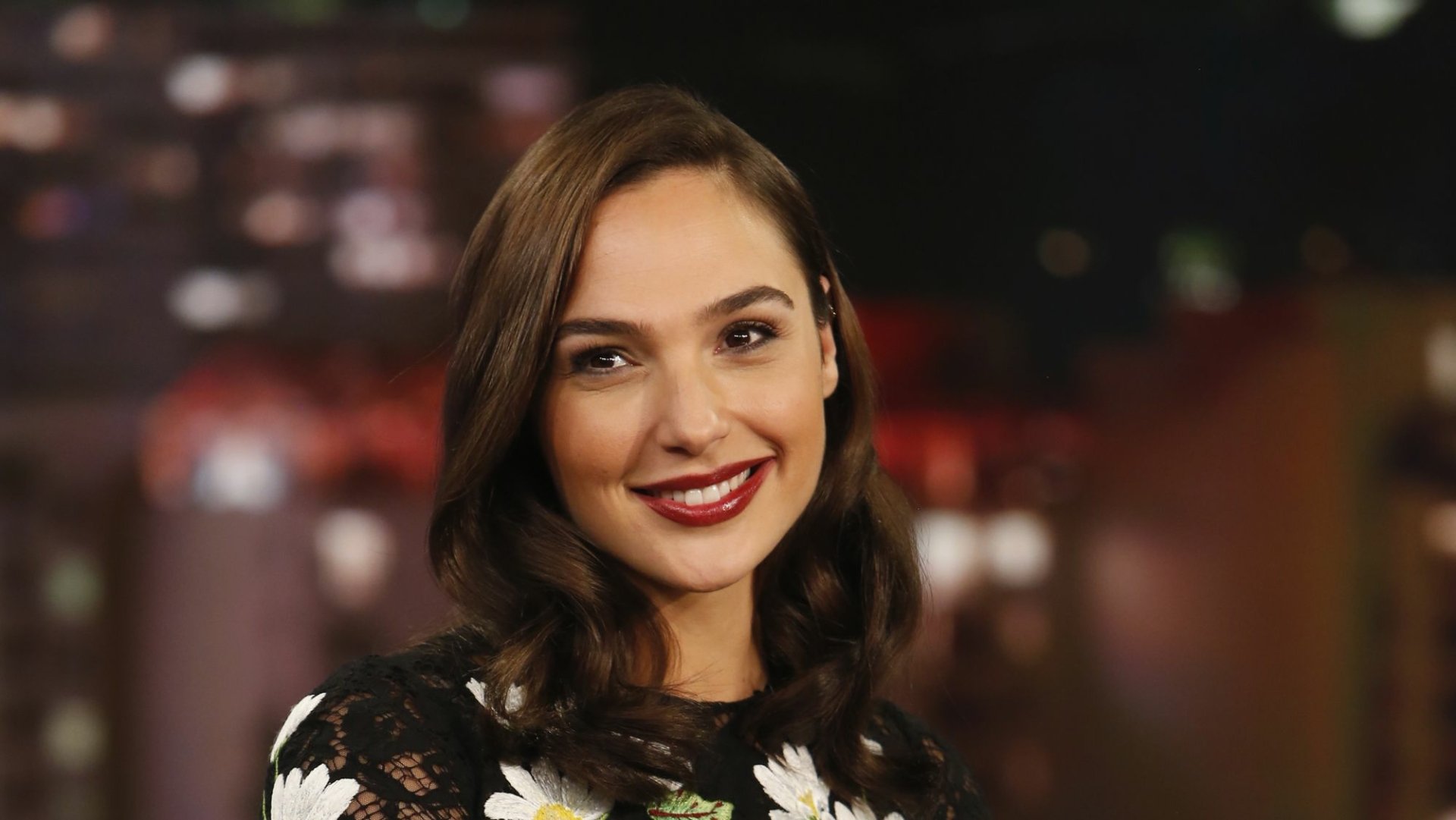 HD PC desktop wallpaper featuring a smiling Gal Gadot with softly curled hair and a floral top against a blurred city night background.