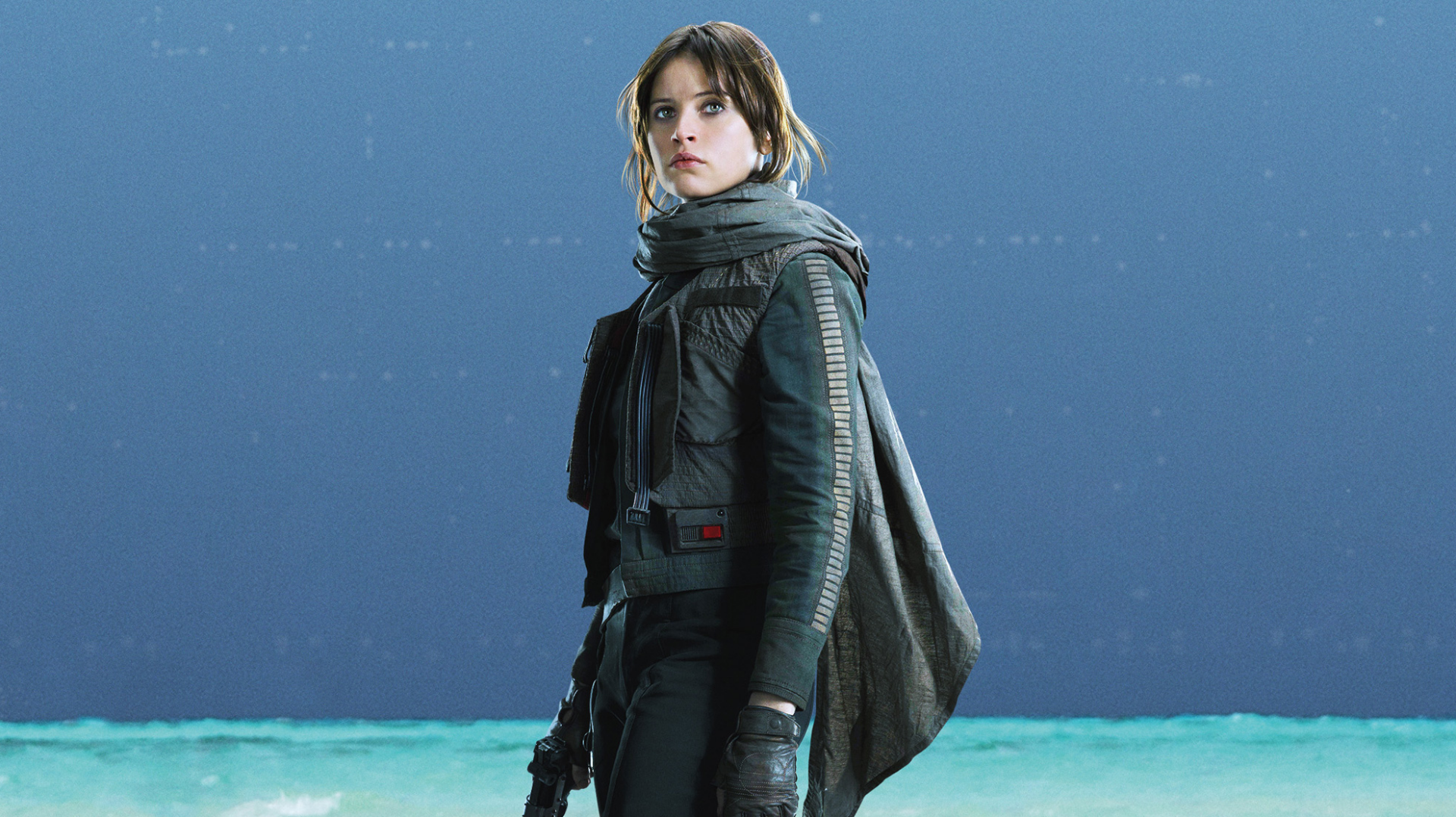 HD desktop wallpaper featuring Felicity Jones as Jyn Erso from the movie Rogue One: A Star Wars Story. She is standing against a serene backdrop of blue ocean and sky.
