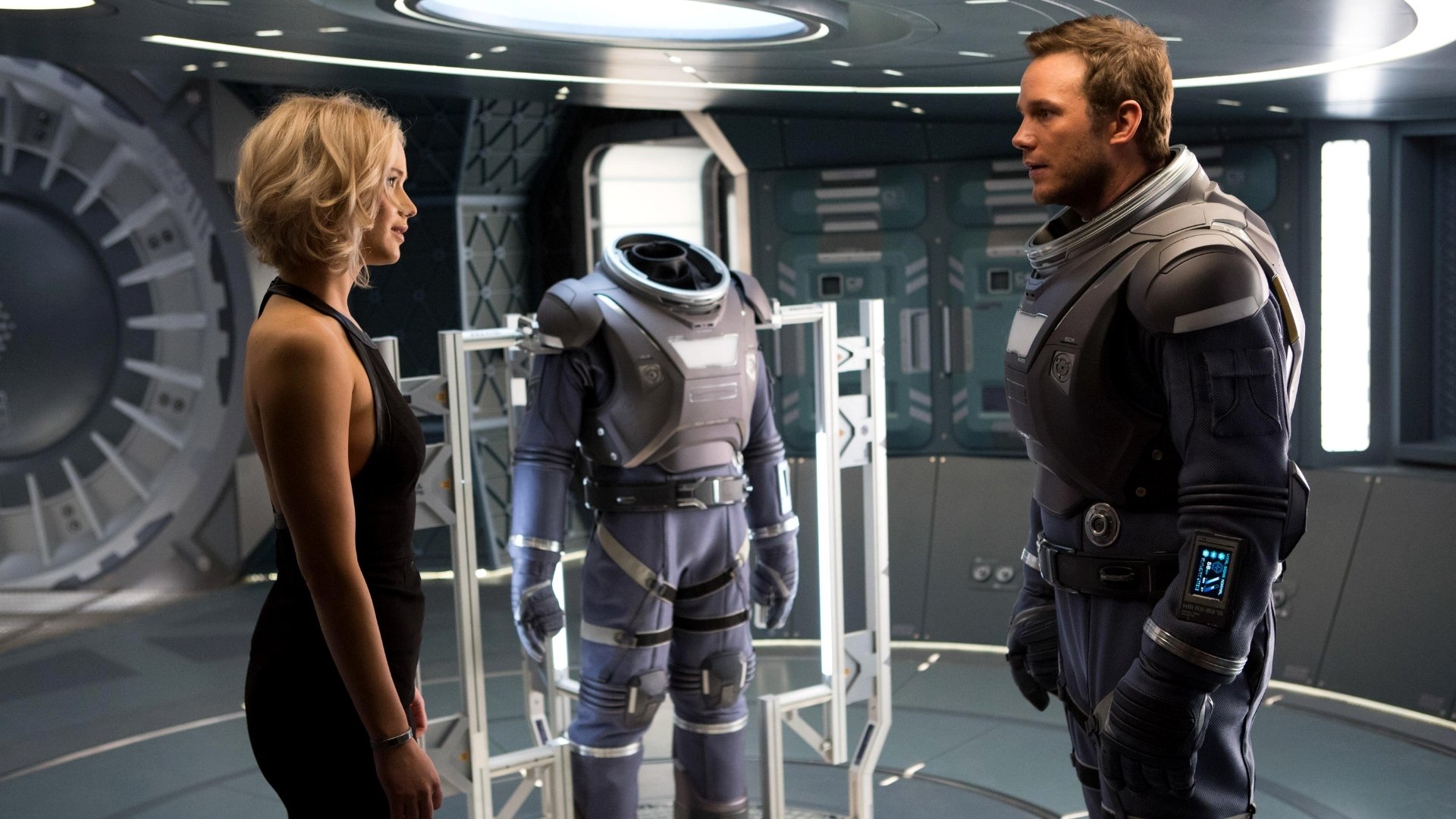 HD PC wallpaper of two actors from the movie Passengers standing in a futuristic spacecraft cabin, a spacesuit displayed between them.