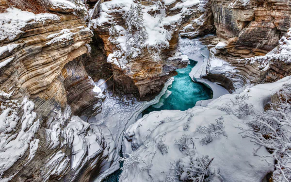 Winter landscape of a frozen turquoise river winding through snow-covered rocky cliffs in Jasper National Park, Canada, showcasing pristine mountain nature.