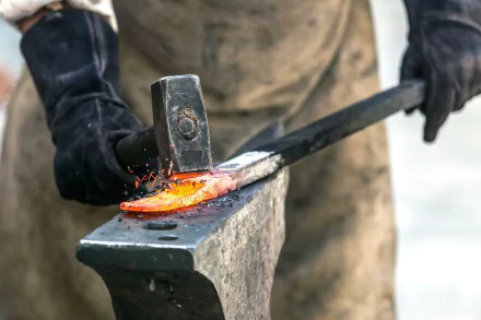 A high-definition desktop wallpaper featuring a blacksmith hammering a glowing piece of metal on an anvil, surrounded by sparks. An intense moment in the smithing process captured in vibrant photography.