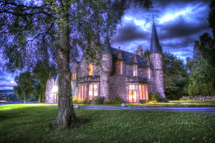 A stunning HDR image of Bunchrew House, a historic castle in Scotland, glowing against a dramatic sky. The building, now a hotel, is surrounded by lush greenery. This HD wallpaper captures its enchanting architecture.