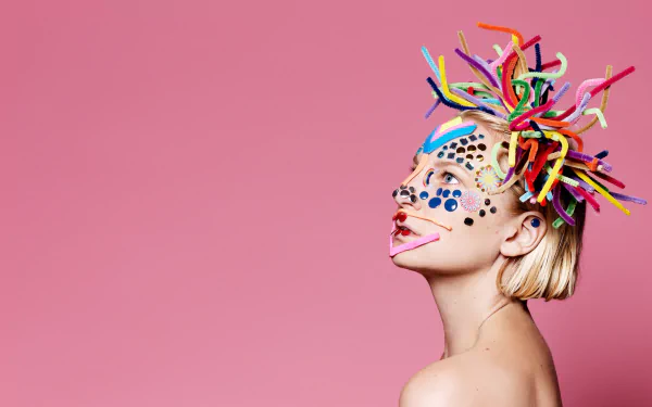 HD desktop wallpaper featuring an artistic portrayal with colorful headpiece and playful face paint on a pink background.