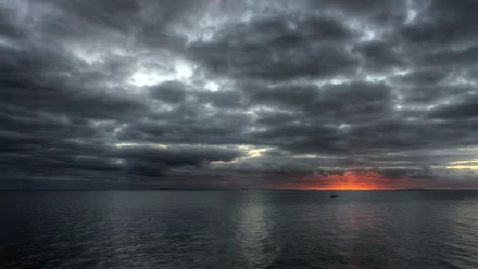 HD desktop wallpaper showing a grey sky filled with clouds over a calm ocean at the horizon, with a hint of orange light breaking through near the water.