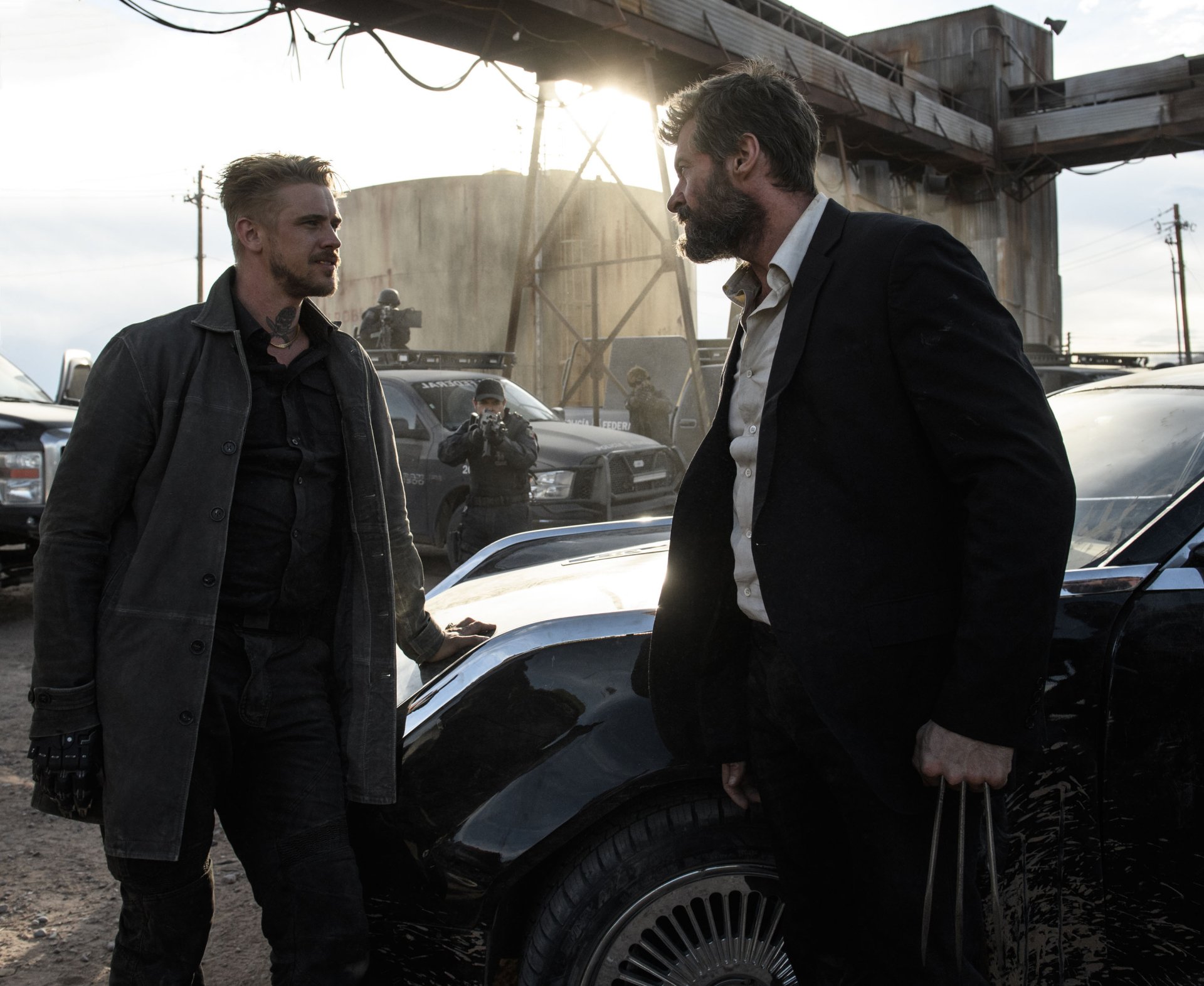 HD wallpaper featuring a scene from the movie Logan with Hugh Jackman engaged in a tense standoff with another character, set in an industrial environment with vehicles and armed personnel in the background.