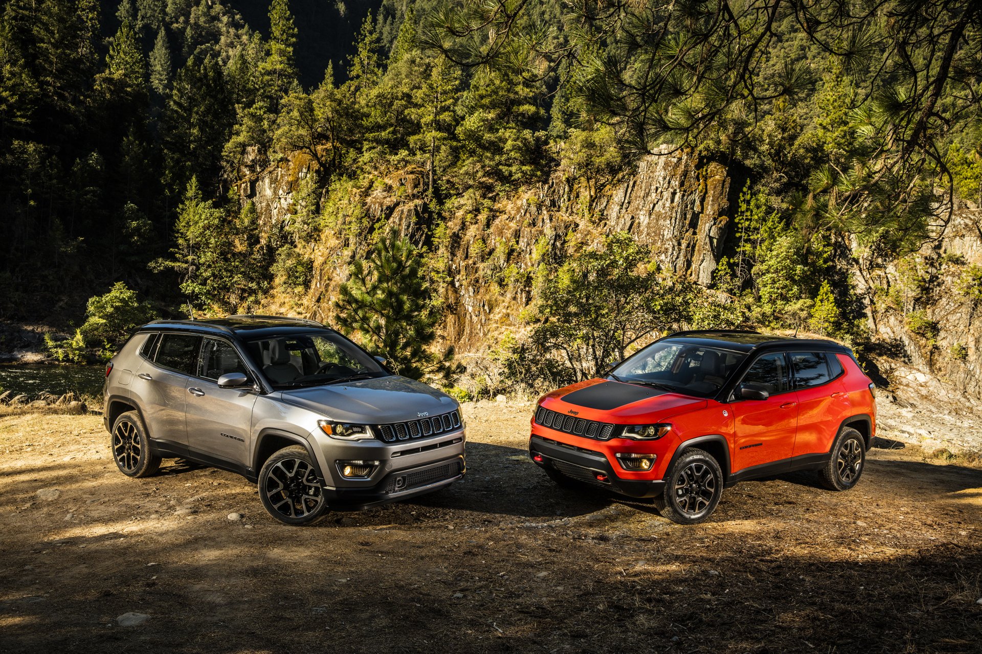 2K Quad HD PC desktop wallpaper of two Jeep Trailhawk SUVs, silver and red, parked on a rugged forest clearing beneath rocky cliffs.