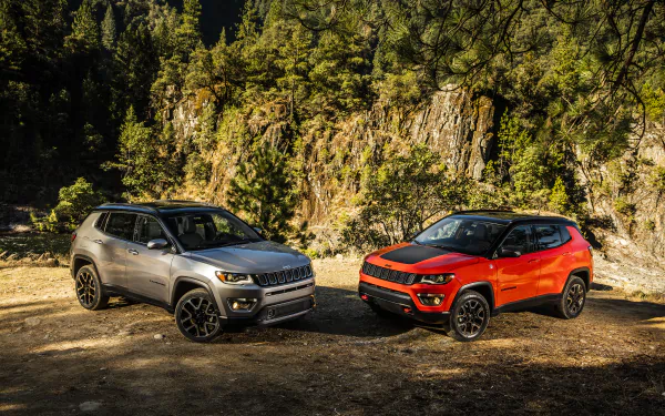 2K Quad HD PC desktop wallpaper of two Jeep Trailhawk SUVs, silver and red, parked on a rugged forest clearing beneath rocky cliffs.
