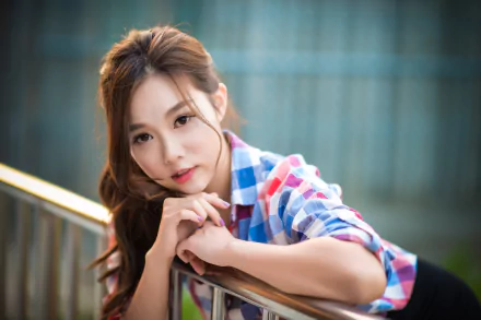 A high-definition desktop wallpaper featuring an Asian brunette model with brown eyes. She is wearing a checkered shirt and posing gracefully against a metallic railing.