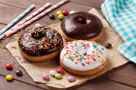 5K Ultra HD PC desktop wallpaper of three colorful doughnuts (chocolate-glazed, sprinkle-topped, white-iced) on a wooden table with candy pieces and striped paper straws — candy, sweets, food, doughnut.