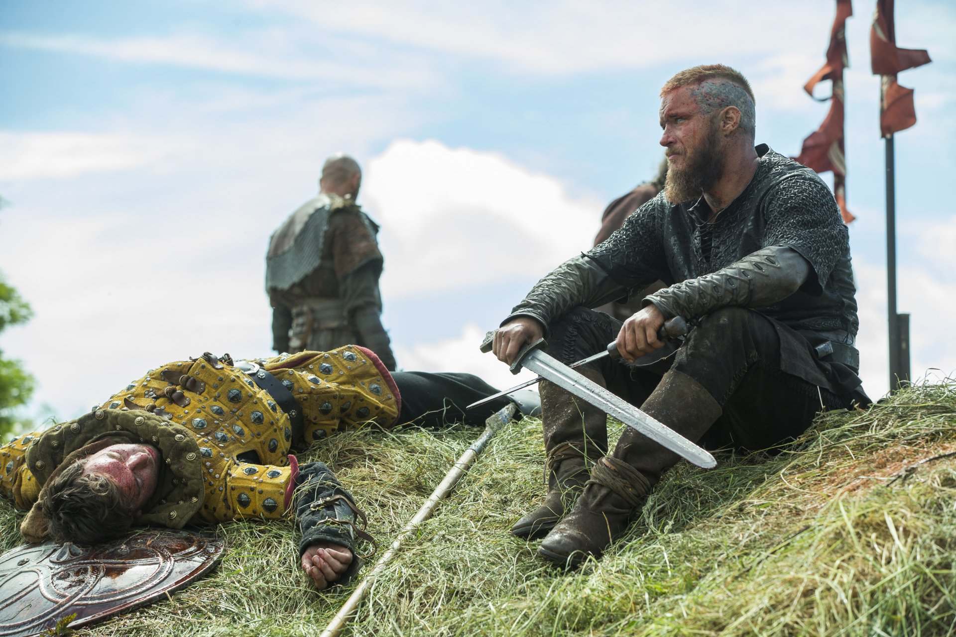 HD PC desktop wallpaper featuring a scene from the TV show Vikings (2013) with a warrior sitting beside a fallen opponent on a grassy hill under a cloudy sky.