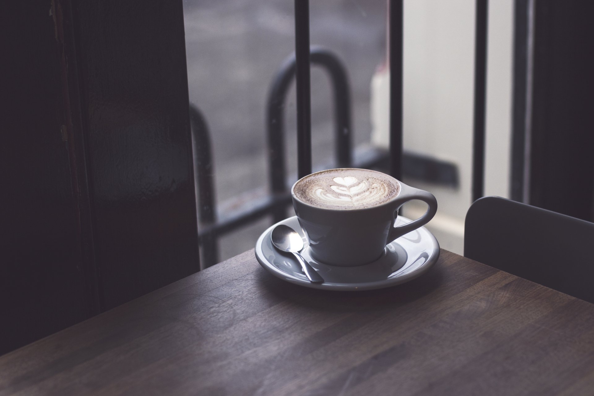 A 4K Ultra HD desktop wallpaper featuring a cup of coffee with latte art on a saucer, placed on a wooden table near a window.