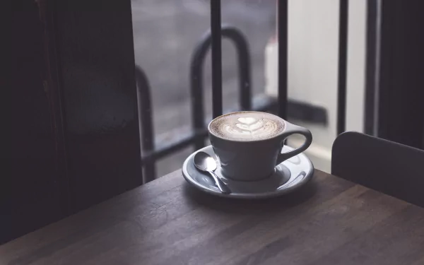 A 4K Ultra HD desktop wallpaper featuring a cup of coffee with latte art on a saucer, placed on a wooden table near a window.