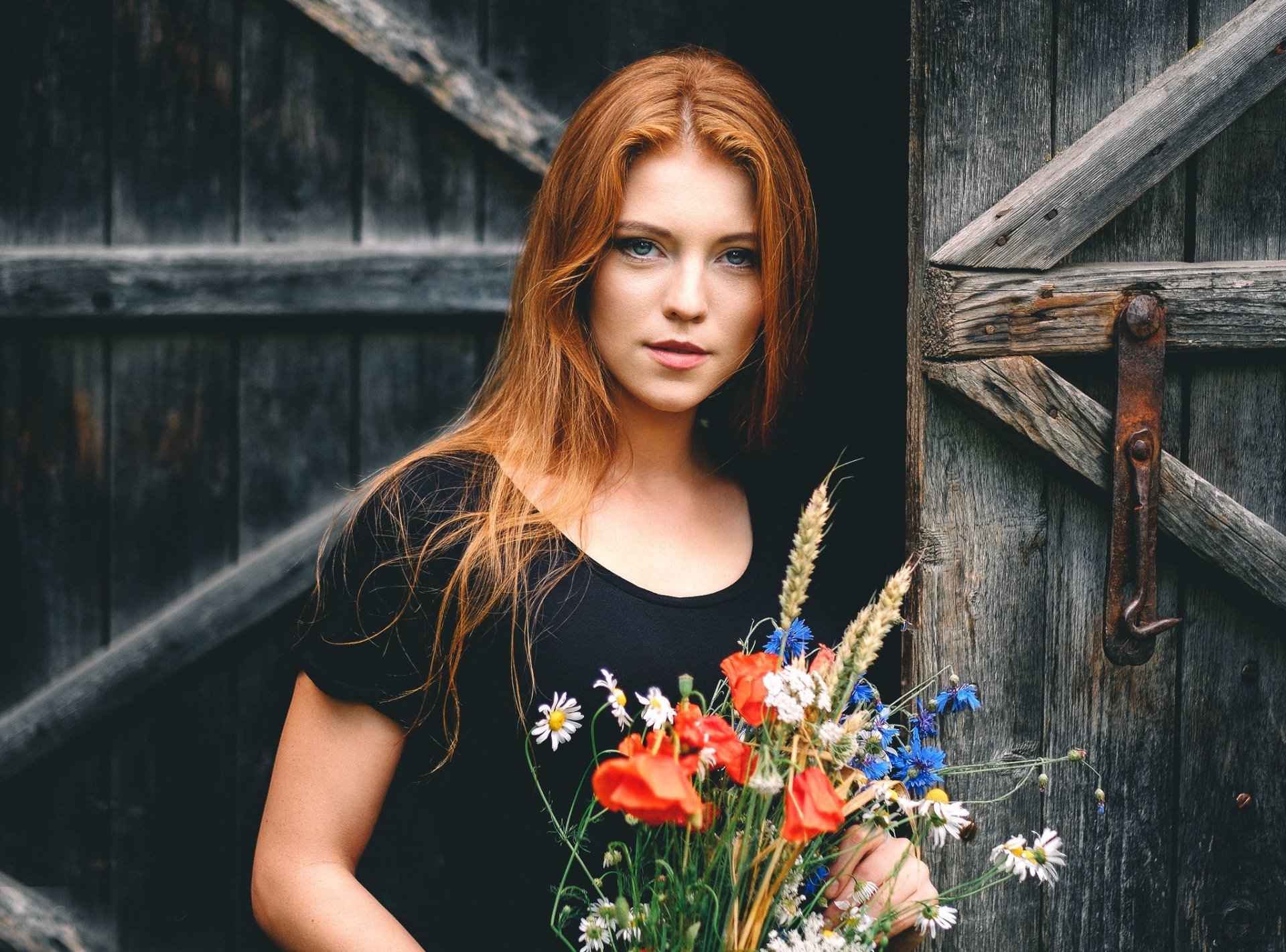 A redheaded woman with blue eyes, dressed in black, holds a bouquet of wildflowers against a rustic wooden background. HD wallpaper and background.