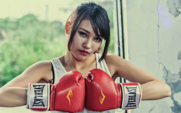 An Asian brunette woman with blue eyes poses confidently in red Everlast boxing gloves against a blurred outdoor background.