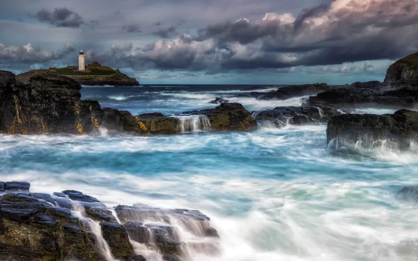 HD desktop wallpaper featuring a foamy ocean with rocks, a distant lighthouse on the horizon, and dramatic clouds overhead. A stunning nature photography scene.
