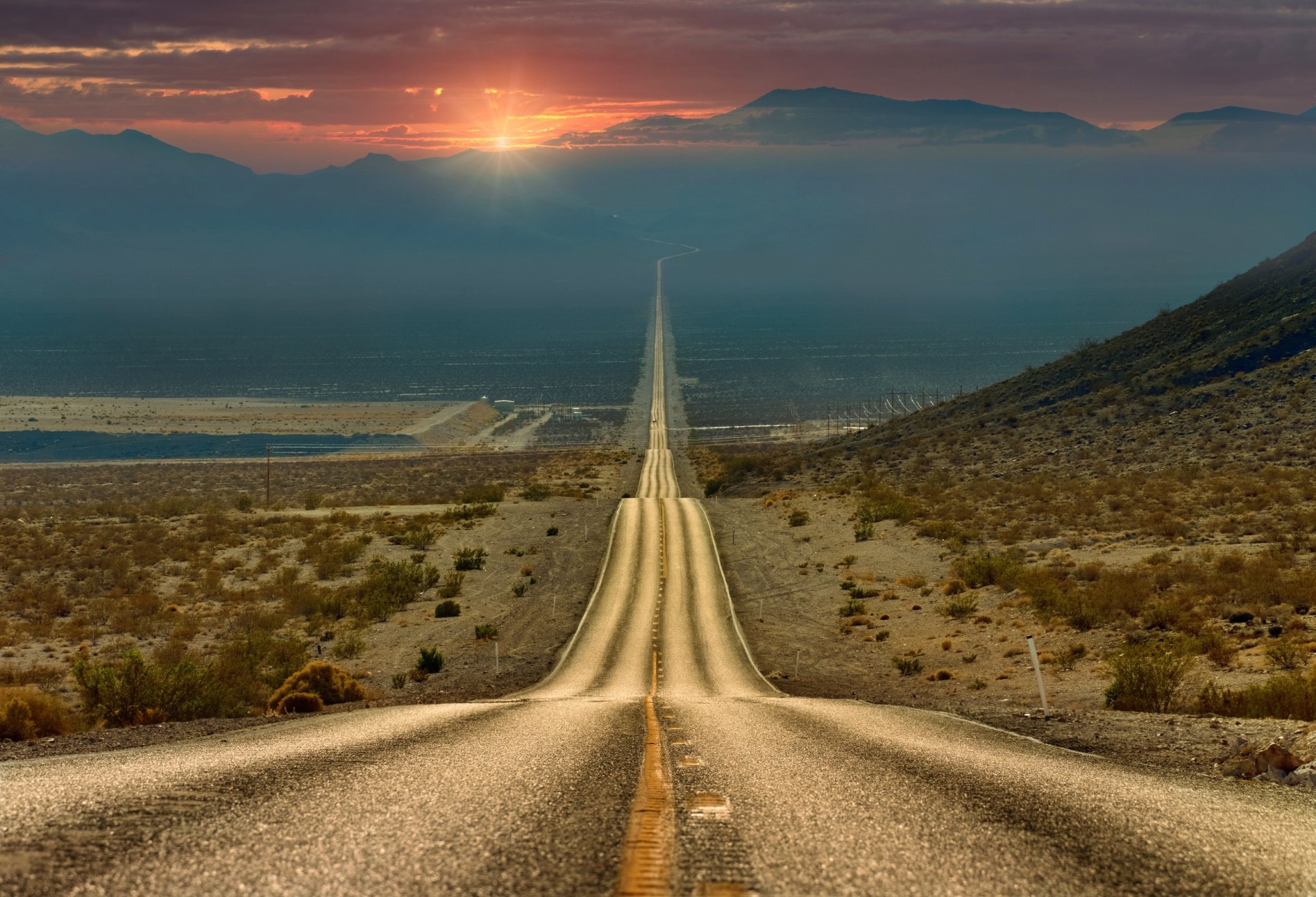 A long, straight road stretches into the sunset across a desert landscape, with mountains in the background. HD desktop wallpaper showcasing nature and man-made beauty.