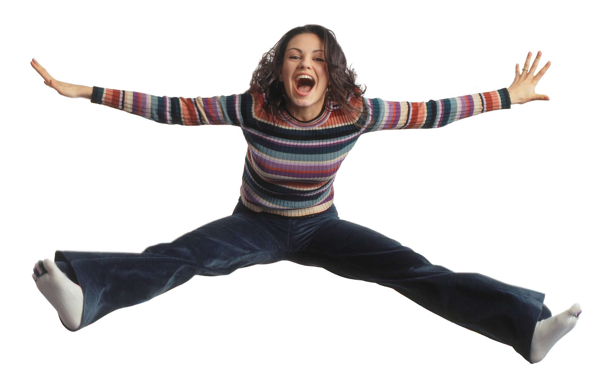 Young woman in a striped sweater and flared jeans, mid-air star jump with arms spread, laughing on white background — HD PC desktop wallpaper with 1970s TV sitcom vibe.