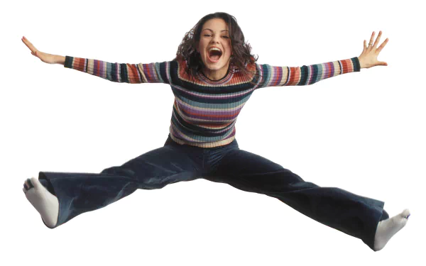 Young woman in a striped sweater and flared jeans, mid-air star jump with arms spread, laughing on white background — HD PC desktop wallpaper with 1970s TV sitcom vibe.