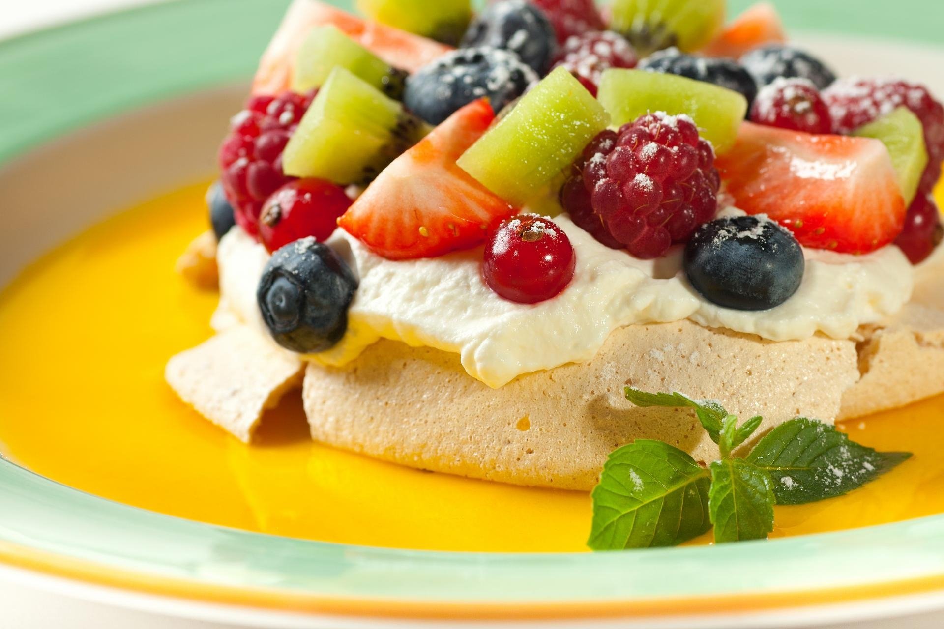 HD desktop wallpaper of a pavlova dessert: crisp meringue topped with whipped cream, strawberries, raspberries, blueberries and kiwi on a pool of yellow sauce, garnished with mint.
