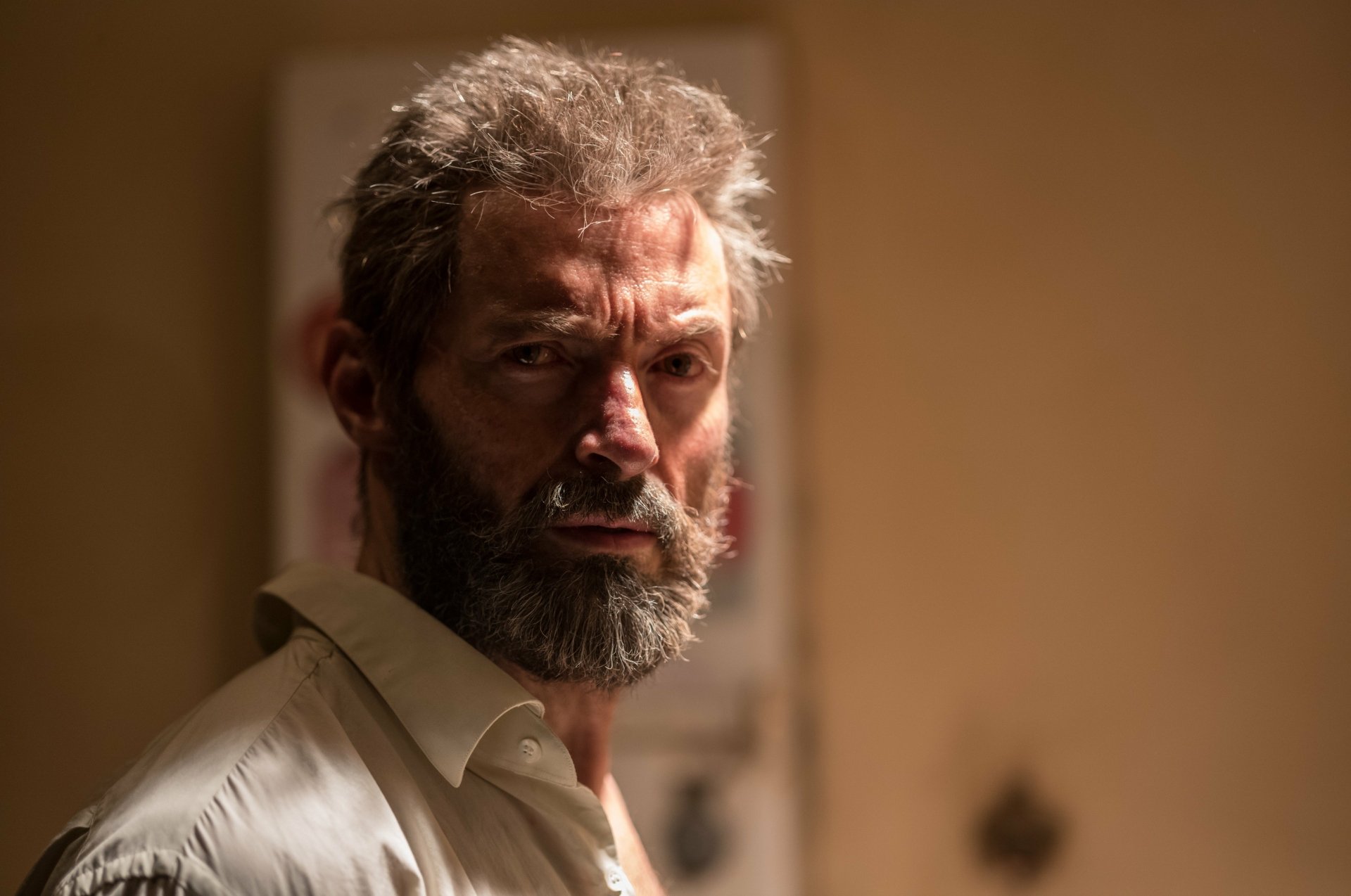 HD desktop wallpaper featuring Hugh Jackman as Wolverine in the movie Logan. Jackman, with a rugged appearance and a serious expression, stands against a dimly lit background.