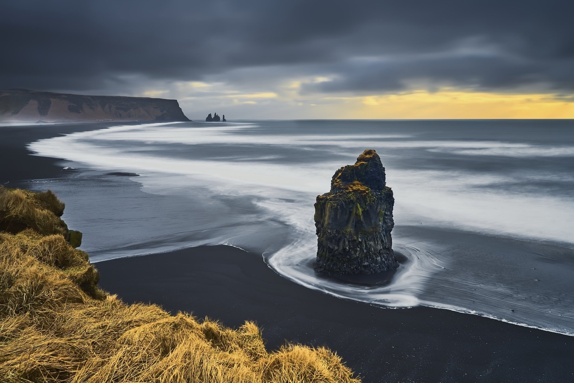 A serene beach scene featuring gentle foam rolling onto a black sand coastline, with dramatic ocean waves meeting a moody horizon under overcast skies.