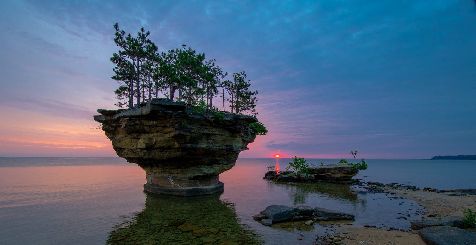 HD desktop wallpaper featuring a breathtaking sunset over the sea, with the sun near the horizon, a tree-topped rock formation in the foreground, and serene natural scenery.