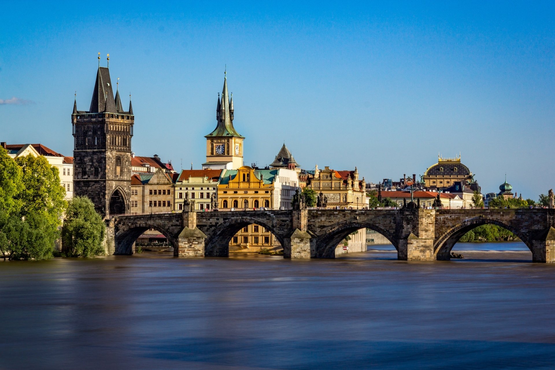 HD PC desktop wallpaper of Prague, Czech Republic: Charles Bridge spanning the Vltava with historic buildings and towers under a clear blue sky.