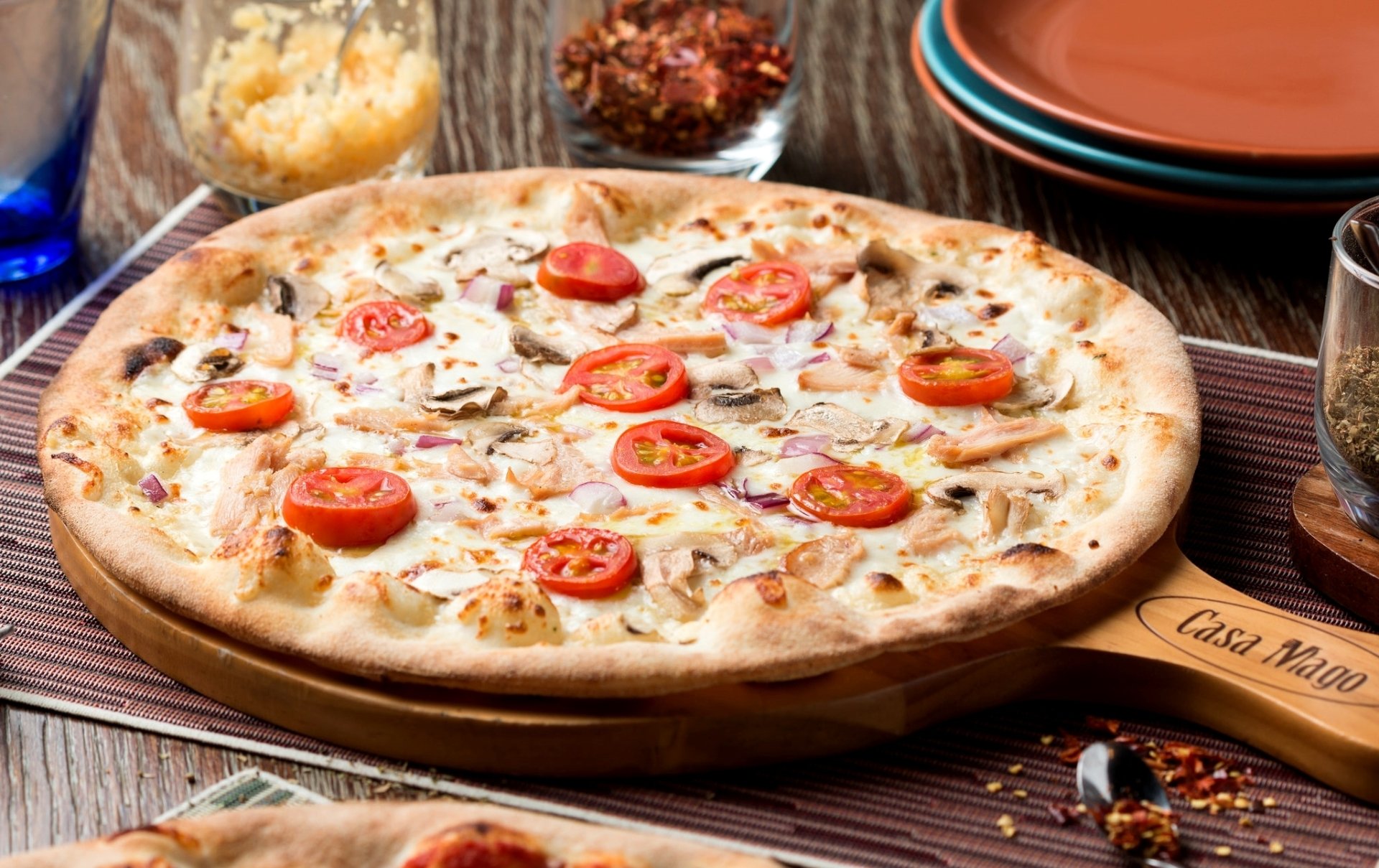 HD PC desktop wallpaper and background: close-up of a hot thin-crust pizza topped with cherry tomatoes, mushrooms and melted cheese on a wooden board with plates and spices nearby.