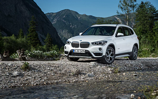 White BMW X1 SUV parked on a rocky riverbank with mountains and trees in the background, captured in 4K Ultra HD as a PC desktop wallpaper and background.