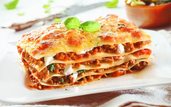 HD desktop wallpaper showing a close-up of a layered lasagna pasta dish garnished with fresh herbs on a white plate.