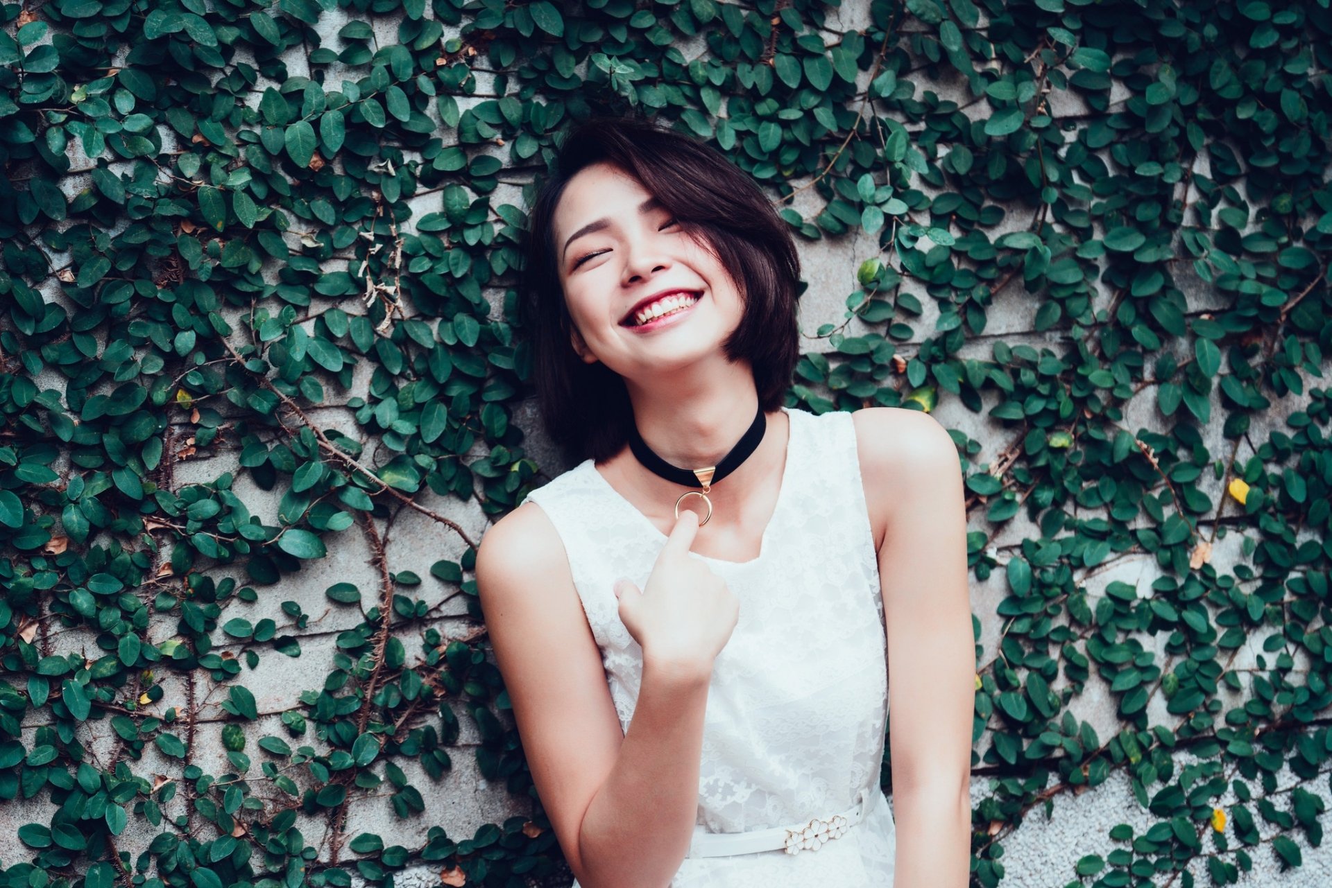 Smiling Asian woman with short brunette hair in a white dress poses against a lush green leafy wall, captured as a vibrant HD desktop wallpaper.