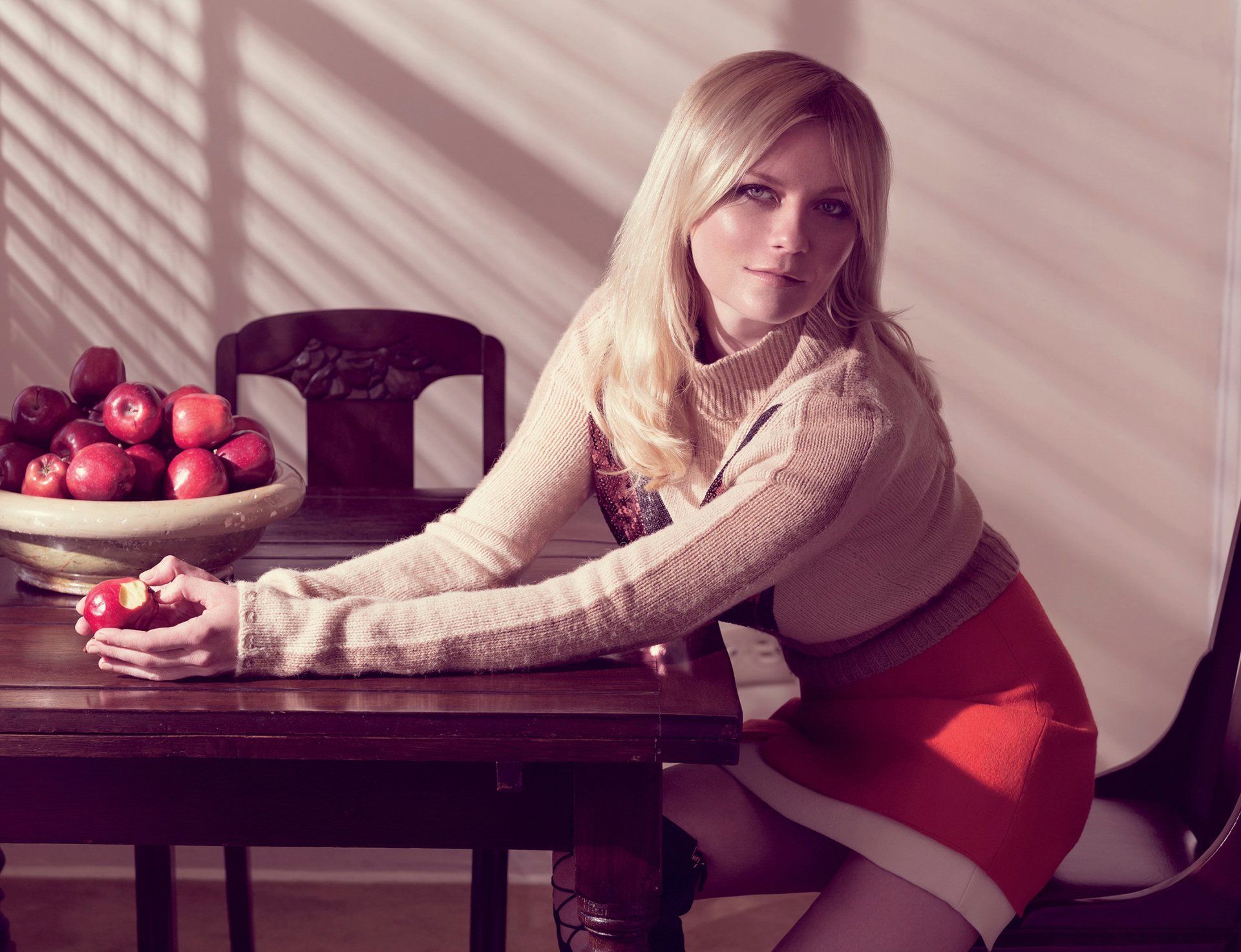 HD wallpaper of American actress with long blonde hair and blue eyes, sitting at a table holding an apple, next to a bowl of red apples in a softly lit room.
