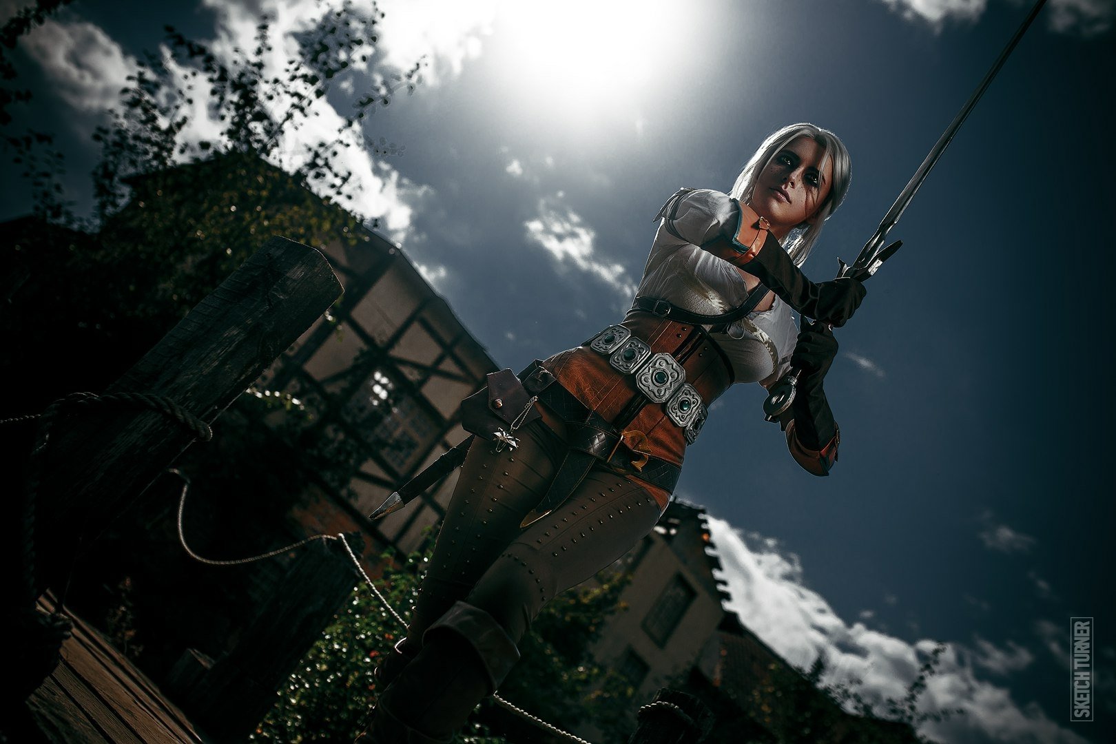 An HD desktop wallpaper featuring a woman cosplaying as Ciri from The Witcher 3: Wild Hunt, posing outdoors with a dramatic sky and sunlight in the background.