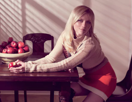 HD wallpaper of American actress with long blonde hair and blue eyes, sitting at a table holding an apple, next to a bowl of red apples in a softly lit room.
