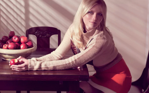 HD wallpaper of American actress with long blonde hair and blue eyes, sitting at a table holding an apple, next to a bowl of red apples in a softly lit room.